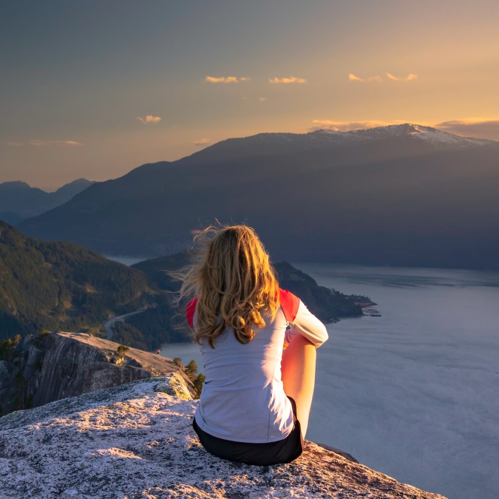 woman in white t-shirt sitting on rock formation near body of water during daytime
